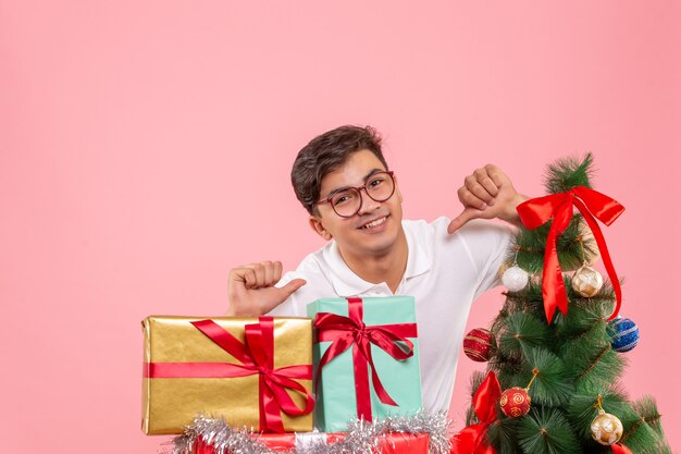 Vista frontal del joven alrededor de regalos de Navidad y árbol de vacaciones en pared rosa