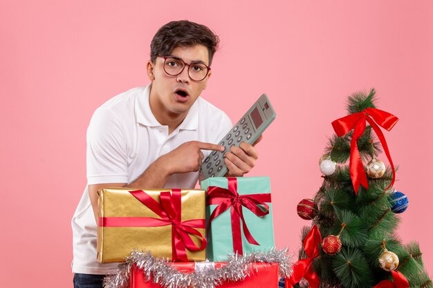 Vista frontal del joven alrededor de regalos de Navidad con calculadora en pared rosa