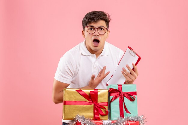 Vista frontal del joven alrededor de regalos de Navidad con nota de archivo confundida en la pared rosa