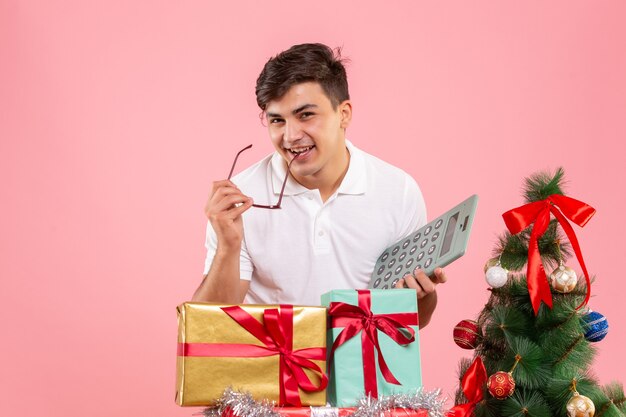 Vista frontal del joven alrededor de regalos de Navidad sosteniendo la calculadora en una pared rosa