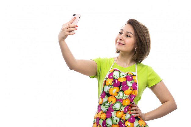 Una vista frontal joven ama de casa hermosa en camisa verde capa colorida sonriendo feliz tomando un selfie en el fondo blanco casa cocina femenina