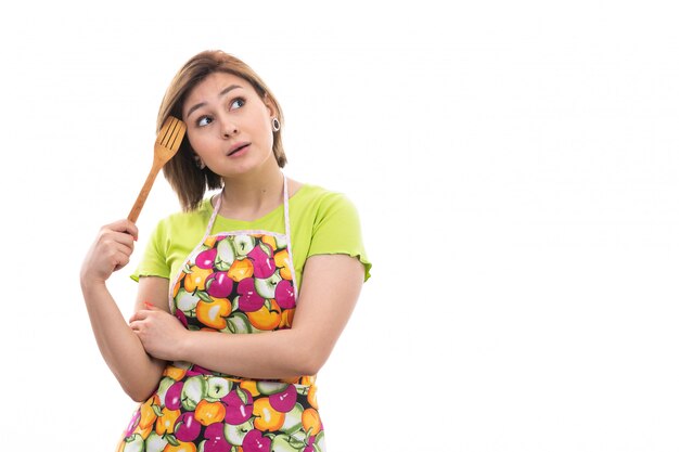 Una vista frontal joven ama de casa hermosa en camisa verde capa colorida sonriendo sosteniendo cocina aparato de madera pensando en el fondo blanco limpieza de la cocina de la casa