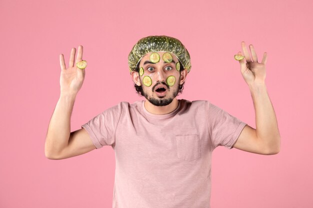 Vista frontal del joven aplicando máscara de pepino en su rostro en la pared rosa