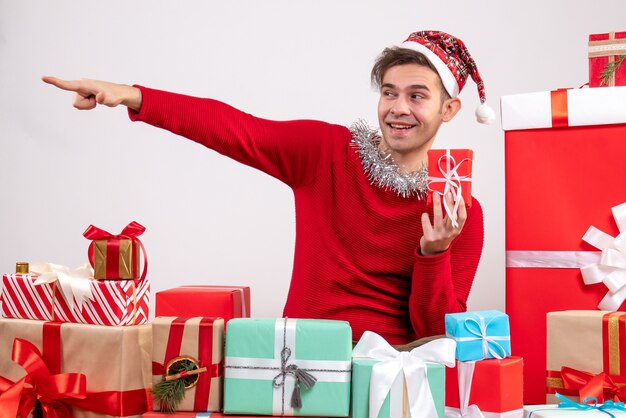 Vista frontal joven apuntando a algo sentado alrededor de regalos de Navidad