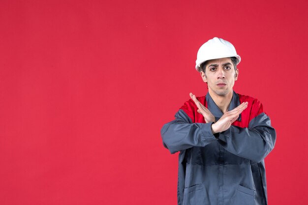 Vista frontal del joven arquitecto nervioso en uniforme con casco y haciendo gesto de parada en la pared roja aislada