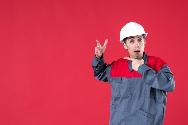 Vista frontal del joven arquitecto sorprendido en uniforme con casco en pared roja aislada