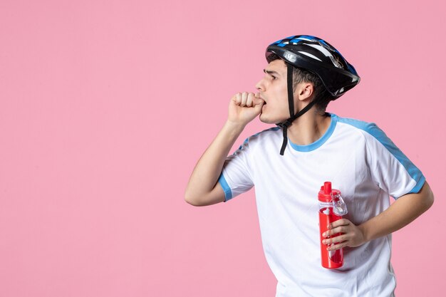 Vista frontal joven atleta masculino en ropa deportiva con casco y botella de agua pared rosa