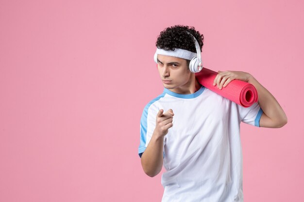 Vista frontal del joven atleta masculino en ropa deportiva con estera de yoga en la pared rosa