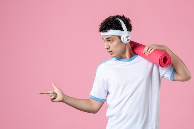 Vista frontal del joven atleta masculino en ropa deportiva con estera de yoga en la pared rosa