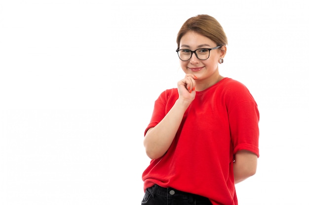 Una vista frontal joven atractiva en camiseta roja con jeans negros en gafas de sol posando sonriente coqueta en el blanco