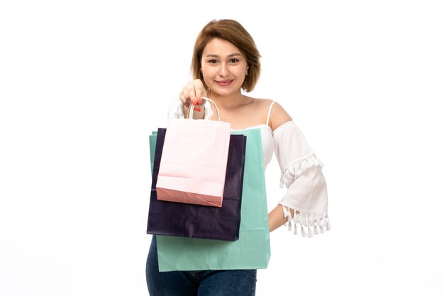 Una vista frontal joven y atractiva dama en camisa blanca y jeans azul con paquetes de compras sonriendo en el blanco