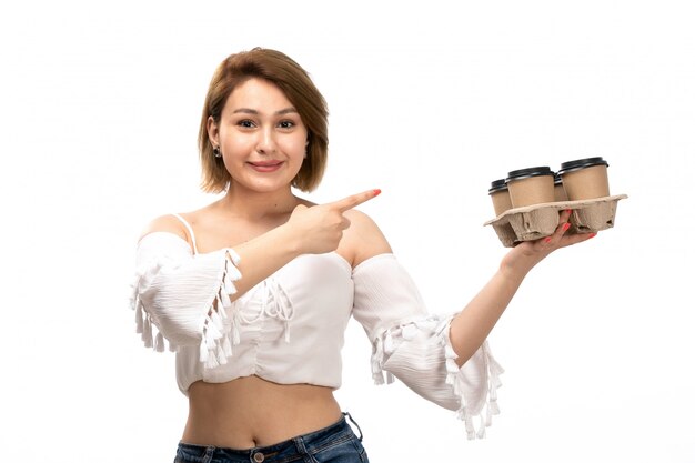 Una vista frontal joven y atractiva dama en camisa blanca y jeans azul sosteniendo tazas de café sonriendo en el blanco