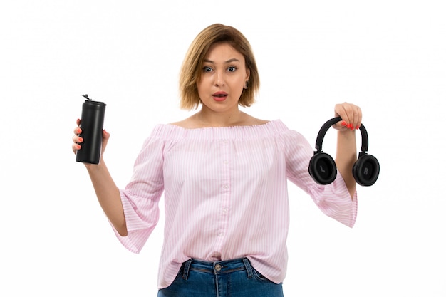 Una vista frontal joven y atractiva dama en camisa rosa y jeans azul con auriculares negros bebiendo con termo negro auriculares negros en el blanco