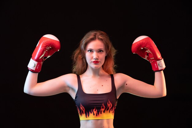 Una vista frontal joven y atractiva dama en guantes de boxeo rojos fuego camisa flexionando sobre el fondo negro entrenamiento de boxeo deportivo