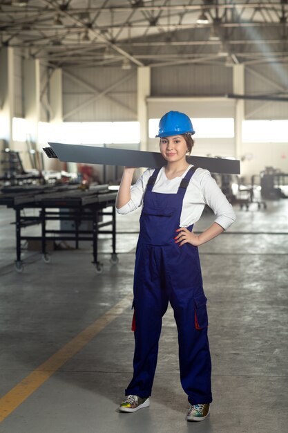 Una vista frontal joven y atractiva dama en traje azul de construcción y casco trabajando sosteniendo algo pesado metálico durante la construcción de arquitectura de edificios diurnos