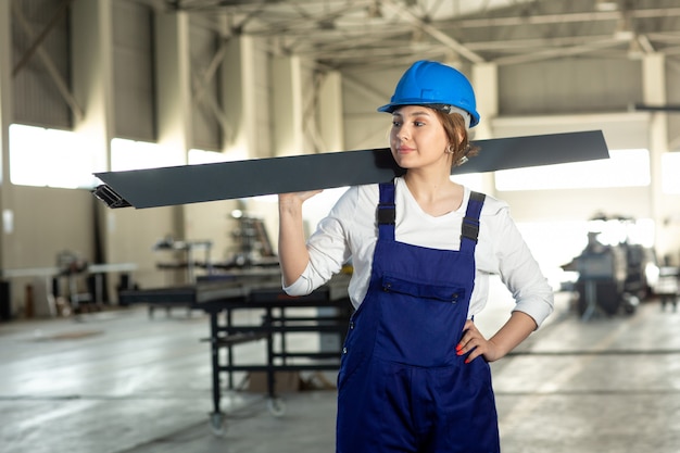 Una vista frontal joven y atractiva dama en traje azul de construcción y casco de trabajo sosteniendo pesados detalles metálicos durante la construcción de arquitectura de edificios durante el día