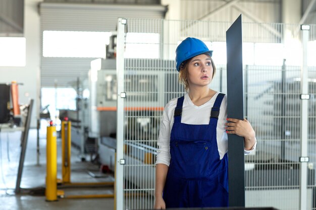 Una vista frontal joven y atractiva dama en traje azul de construcción y casco de trabajo sosteniendo pesados detalles metálicos durante la construcción de arquitectura de edificios durante el día