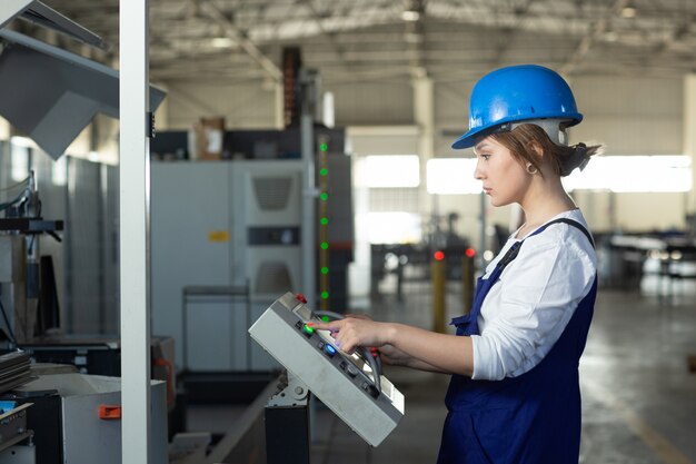 Una vista frontal joven y atractiva dama en traje azul de construcción y máquinas de control de casco en hangar trabajando durante la construcción de arquitectura de edificios diurnos