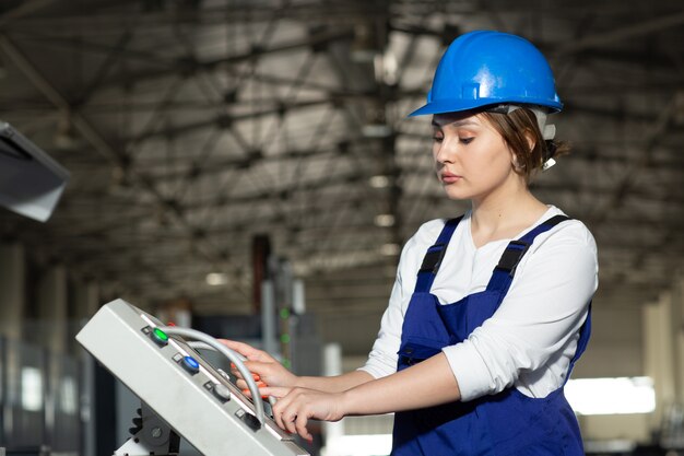 Una vista frontal joven y atractiva dama en traje azul de construcción y máquinas de control de casco en hangar trabajando durante la construcción de arquitectura de edificios diurnos