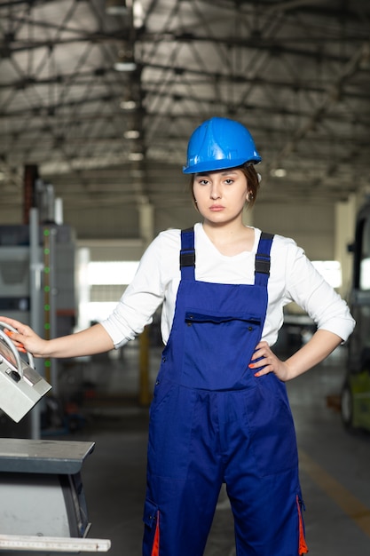 Una vista frontal joven y atractiva dama en traje azul de construcción y máquinas de control de casco en hangar trabajando durante la construcción de arquitectura de edificios diurnos