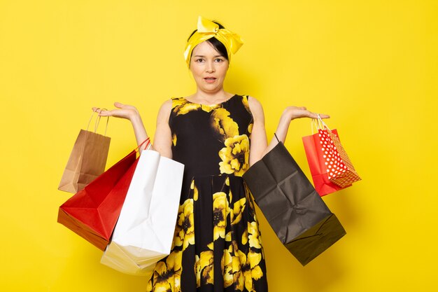 Una vista frontal joven y atractiva dama en vestido amarillo-negro diseñado con flores con vendaje amarillo en la cabeza posando sosteniendo paquetes de compras en el amarillo