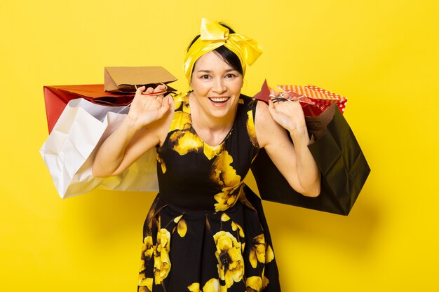 Una vista frontal joven y atractiva dama en vestido amarillo-negro diseñado con flores con vendaje amarillo en la cabeza posando sosteniendo paquetes de compras en el amarillo