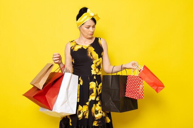 Una vista frontal joven y atractiva dama en vestido amarillo-negro diseñado con flores con vendaje amarillo en la cabeza posando sosteniendo paquetes de compras en el amarillo