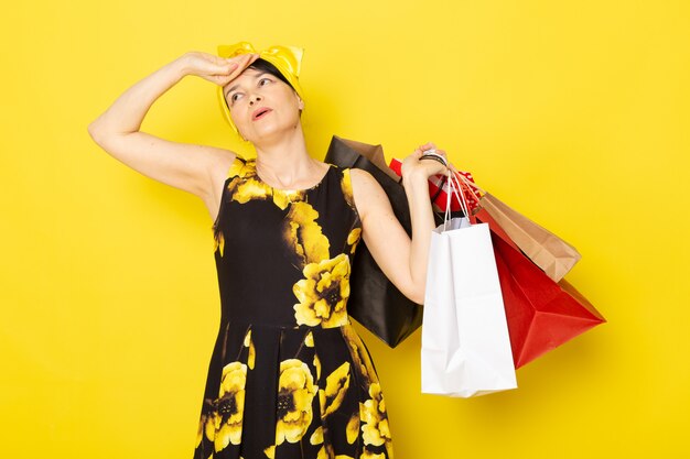 Una vista frontal joven y atractiva dama en vestido de flores amarillo-negro diseñado con vendaje amarillo en la cabeza cansada sosteniendo paquetes de compras en el amarillo