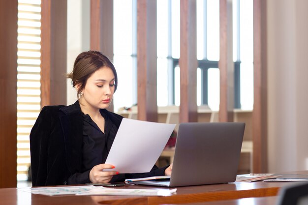 Una vista frontal joven y atractiva empresaria en camisa negra chaqueta negra usando su computadora portátil plateada leyendo leyendo trabajando dentro de su trabajo de oficina edificio de trabajo