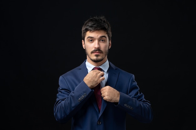 Vista frontal del joven barbudo en traje enderezando su corbata en la pared oscura