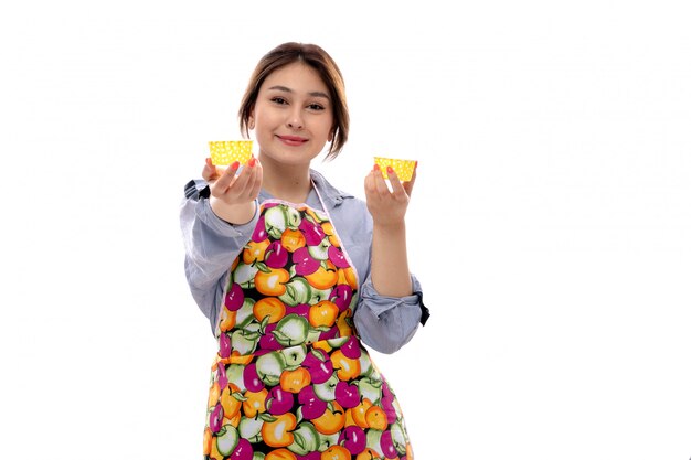 Una vista frontal joven y bella dama en camisa azul claro y capa colorida con moldes para pasteles amarillos sonriendo feliz