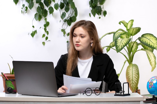 Una vista frontal joven bella dama en camisa blanca y chaqueta negra trabajando con documentos usando su computadora portátil frente a la mesa con hojas colgando