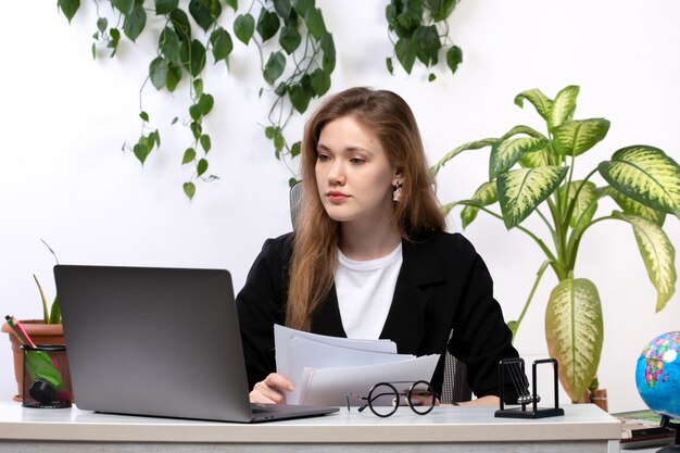 Una vista frontal joven bella dama en camisa blanca y chaqueta negra trabajando con documentos usando su computadora portátil frente a la mesa con hojas colgando