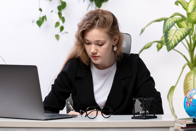 Una vista frontal joven y bella dama en camisa blanca y chaqueta negra usando su computadora portátil en frente de la mesa con hojas colgando