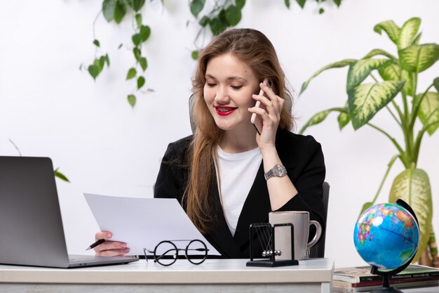 Una vista frontal joven y bella dama en camisa blanca y chaqueta negra usando su computadora portátil frente a la mesa sonriendo hablando por teléfono trabajando con documentos