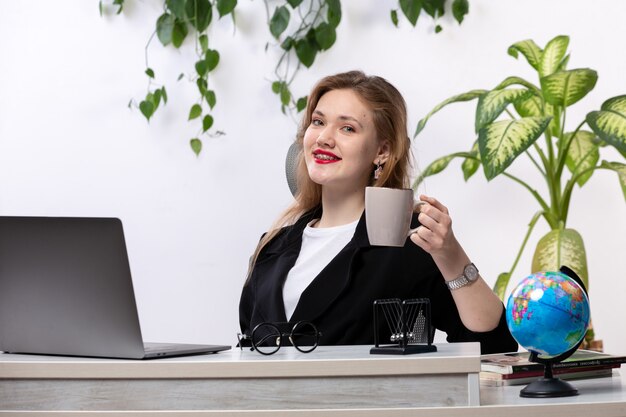Una vista frontal joven bella dama en camisa blanca y chaqueta negra usando su computadora portátil en frente de la mesa sonriendo sosteniendo la taza con hojas colgando