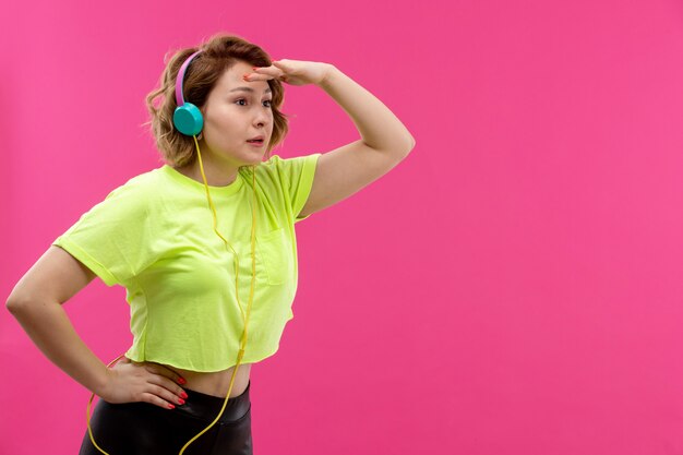 Una vista frontal joven y bella dama en camisa de color ácido pantalón negro con auriculares azules escuchando música mirando a lo lejos