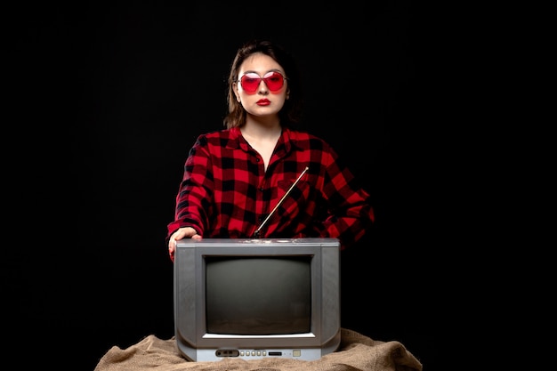 Foto gratuita una vista frontal joven bella dama en camisa a cuadros rojo-negro en gafas de sol rojas cerca de little tv
