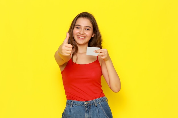 Una vista frontal joven y bella dama en camisa roja y jeans azul sonriendo y sosteniendo la tarjeta