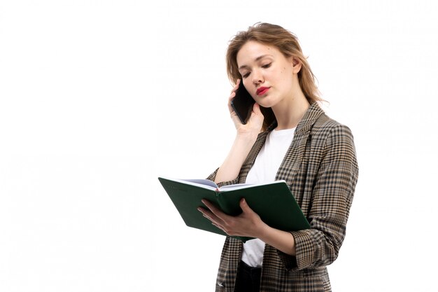 Una vista frontal joven bella dama en camiseta blanca jeans negros y abrigo con smartphone negro hablando y libro verde sobre el blanco