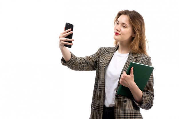 Una vista frontal joven bella dama en camiseta blanca jeans negros y abrigo con smartphone negro tomando un selfie y libro verde sobre el blanco