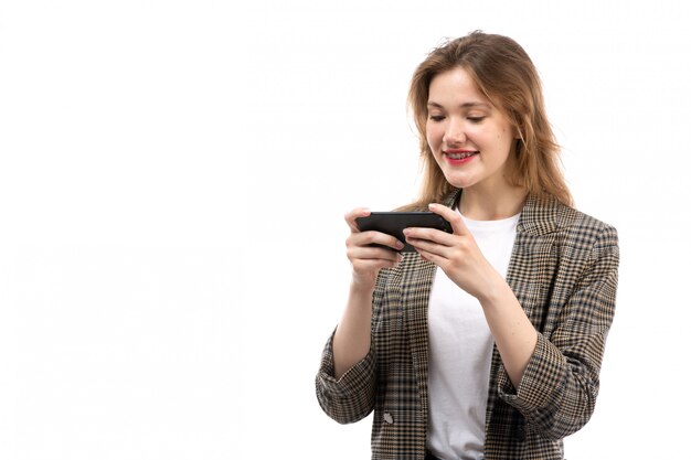 Una vista frontal joven bella dama en camiseta blanca jeans negros y abrigo con teléfono inteligente sonriendo en el blanco