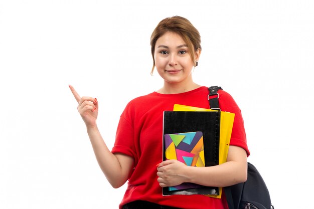 Una vista frontal joven y bella dama en camiseta roja jeans negros con diferentes cuadernos y archivos sonriendo con bolsa en el blanco