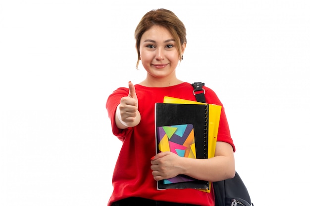 Una vista frontal joven y bella dama en camiseta roja jeans negros con diferentes cuadernos y archivos sonriendo como cartel con bolsa en el blanco