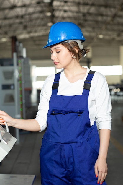 Una vista frontal joven y bella dama en traje de construcción azul y máquinas de control de casco en hangar trabajando durante la construcción de arquitectura de edificios diurnos