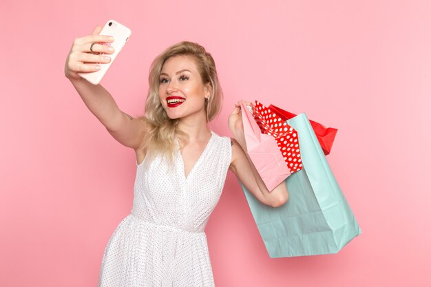 Una vista frontal joven y bella dama en vestido blanco con paquetes de compras con una sonrisa en su rostro tomando un selfie ropa de moda femenina