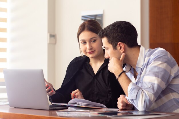 Una vista frontal joven y bella empresaria en camisa negra chaqueta negra junto con un joven usando su computadora portátil plateada discutiendo temas dentro de su trabajo de oficina