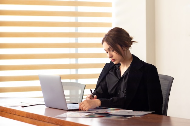 Una vista frontal joven y bella empresaria en camisa negra chaqueta negra usando su computadora portátil plateada leyendo leyendo trabajando dentro de su trabajo de oficina edificio de trabajo