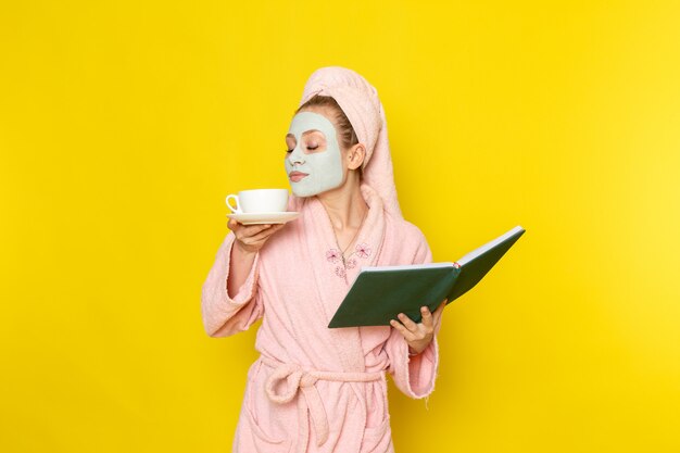 Una vista frontal joven y bella mujer en bata de baño rosa con libro verde y una taza de té