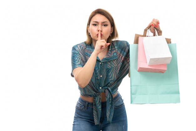 Una vista frontal joven y bella mujer en blusa de color azul jeans sosteniendo paquetes de compras mostrando señal de silencio